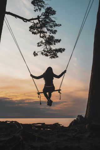 A woman swings on a tree above the sea at sunset, creating a serene silhouette.