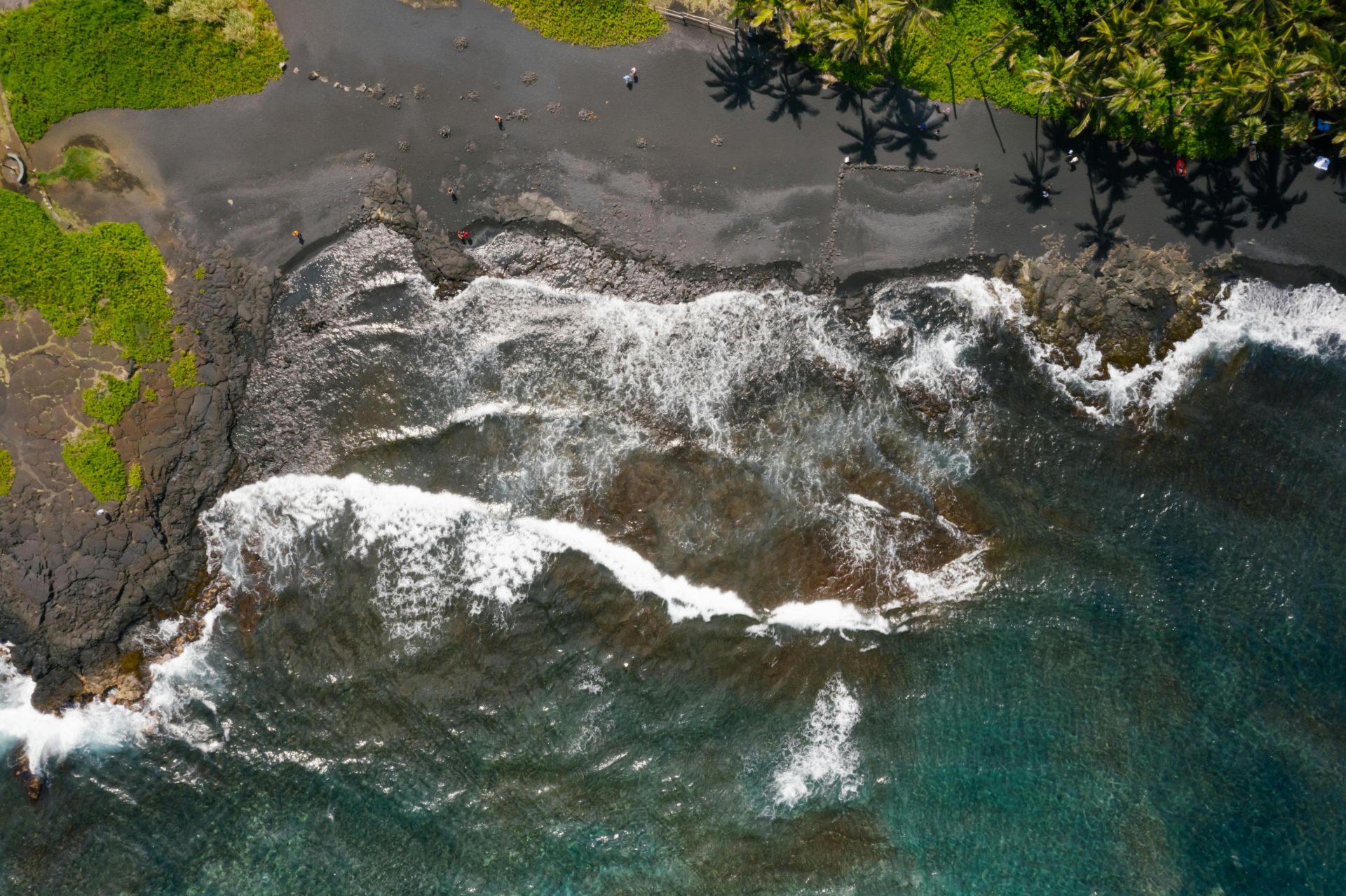 Stunning aerial view of a black sand beach with waves crashing against the shore and lush green palm trees.
