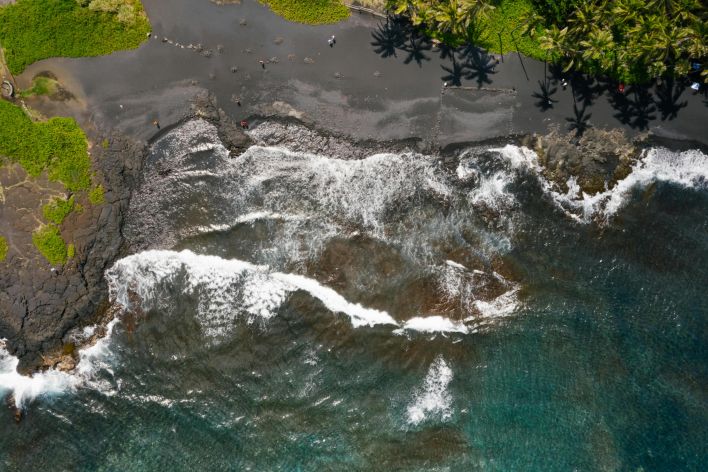 Stunning aerial view of a black sand beach with waves crashing against the shore and lush green palm trees.