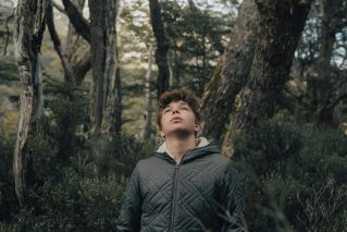 Teen boy wearing jacket, looking up among trees in a forest, representing adventure and exploration.